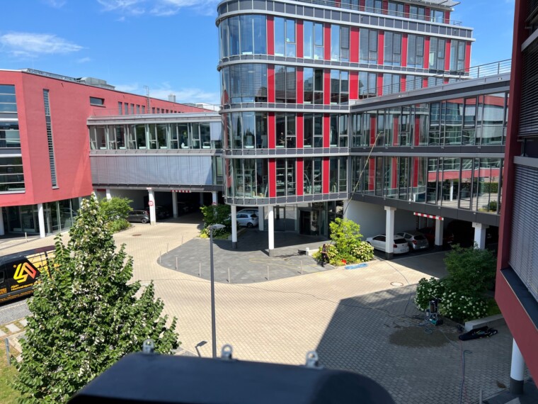 Gewerbegebäude mit mehrgeschossiger Glasfassade und verglasten Verbindungen; im Innenhof sind Mitarbeiter, Teleskoptechnik und ein Alpin-Fahrzeug zu sehen.