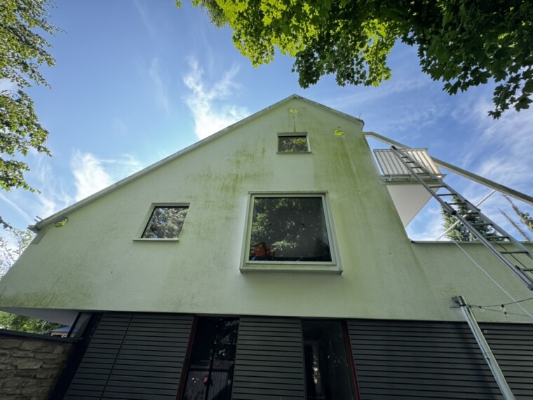 Helle Giebelfassade mit grünlichen Laufspuren, mehreren Fenstern und einer Leiter am rechten Balkon.