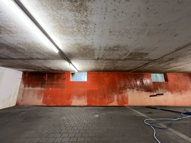 Tiefgarage mit dunklen Flecken an der Decke, orangefarbener Rückwand und Pflasterboden.