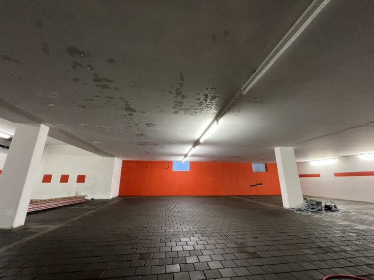 Weitwinkelansicht einer Tiefgarage mit orangefarbener Rückwand, weißen Stützen, Pflasterboden und Reinigungsgeräten.