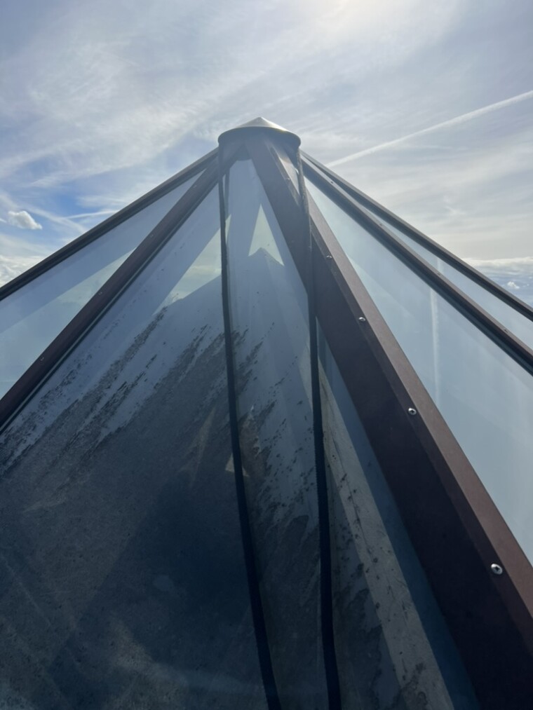 Nahaufnahme einer pyramidenförmigen Dachverglasung mit dunklen, streifenförmigen Belägen auf mehreren Glasscheiben und braunen Metallprofilen.