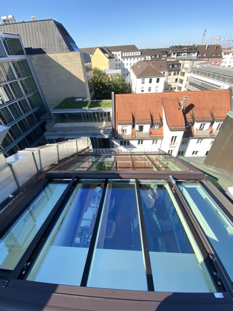 Blick von oben auf eine Dachverglasung mit braunen Profilen, Spiegelungen und sichtbarem Innenraum; im Hintergrund stehen Stadtgebäude.