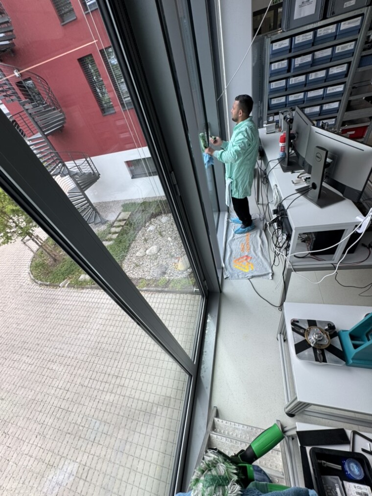 Mitarbeiter in grünem Schutzkittel und Schuhüberziehern bearbeitet eine raumhohe Glasfläche mit einem Handwerkzeug und einem Tuch.