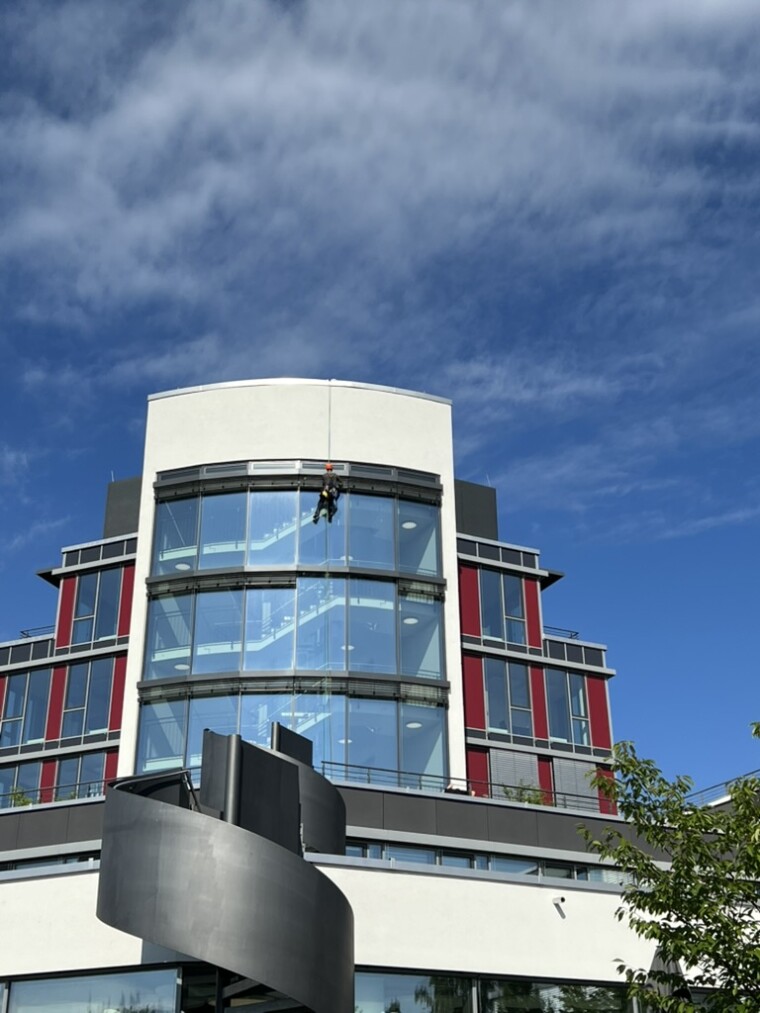 An Seilen hängender Mitarbeiter vor der hohen Glasfront eines Gebäudes mit roten und grauen Fassadenelementen.