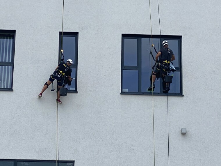 Zwei Mitarbeiter mit Seilausrüstung reinigen außenliegende Fenster an einer hellen Gebäudefassade.